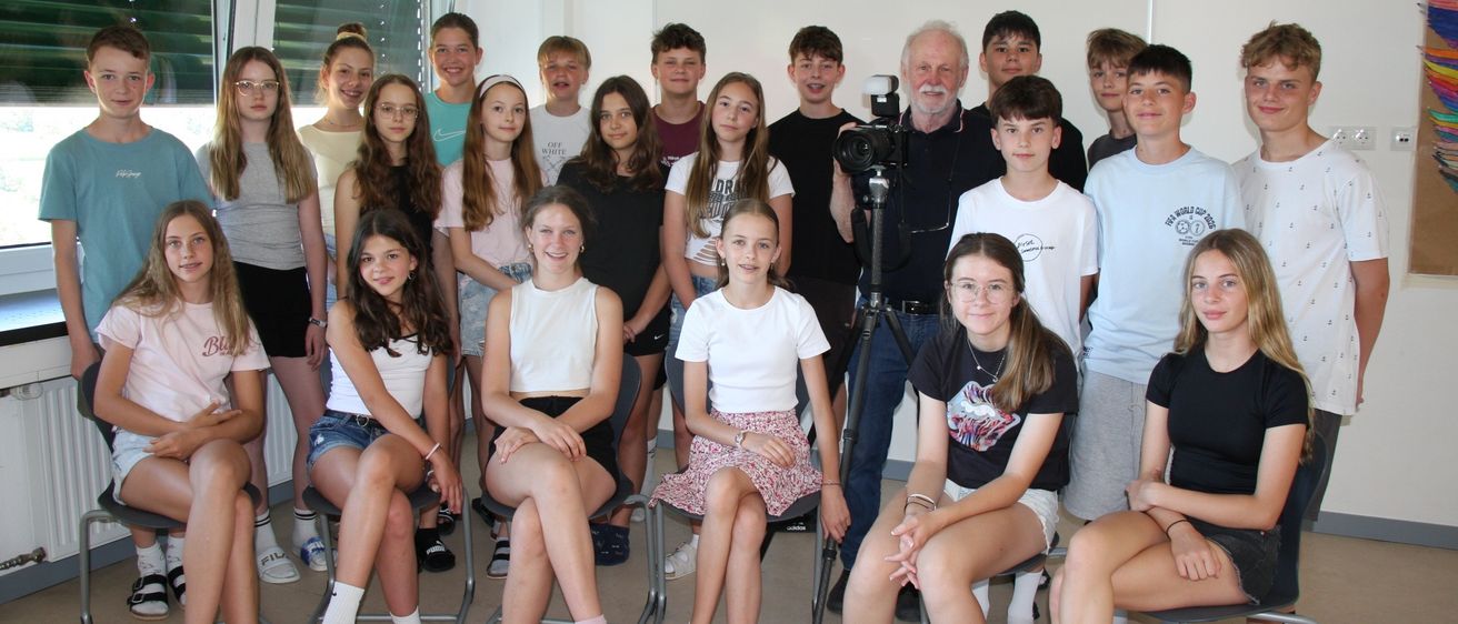 Eine Gruppe junger Menschen und ein Mann versammeln sich in einem Raum, möglicherweise für ein Fotoshooting. Sie sitzen auf Stühlen, ein Mann steht hinter einer Kamera mit Stativ. Der Raum hat weiße Wände und Fenster mit Jalousien.