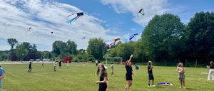 Eine Gruppe von Menschen fliegt Drachen in einem Grasfeld unter einem blauen Himmel mit verstreuten Wolken. Bäume und ein Fußballtor sind im Hintergrund zu sehen.