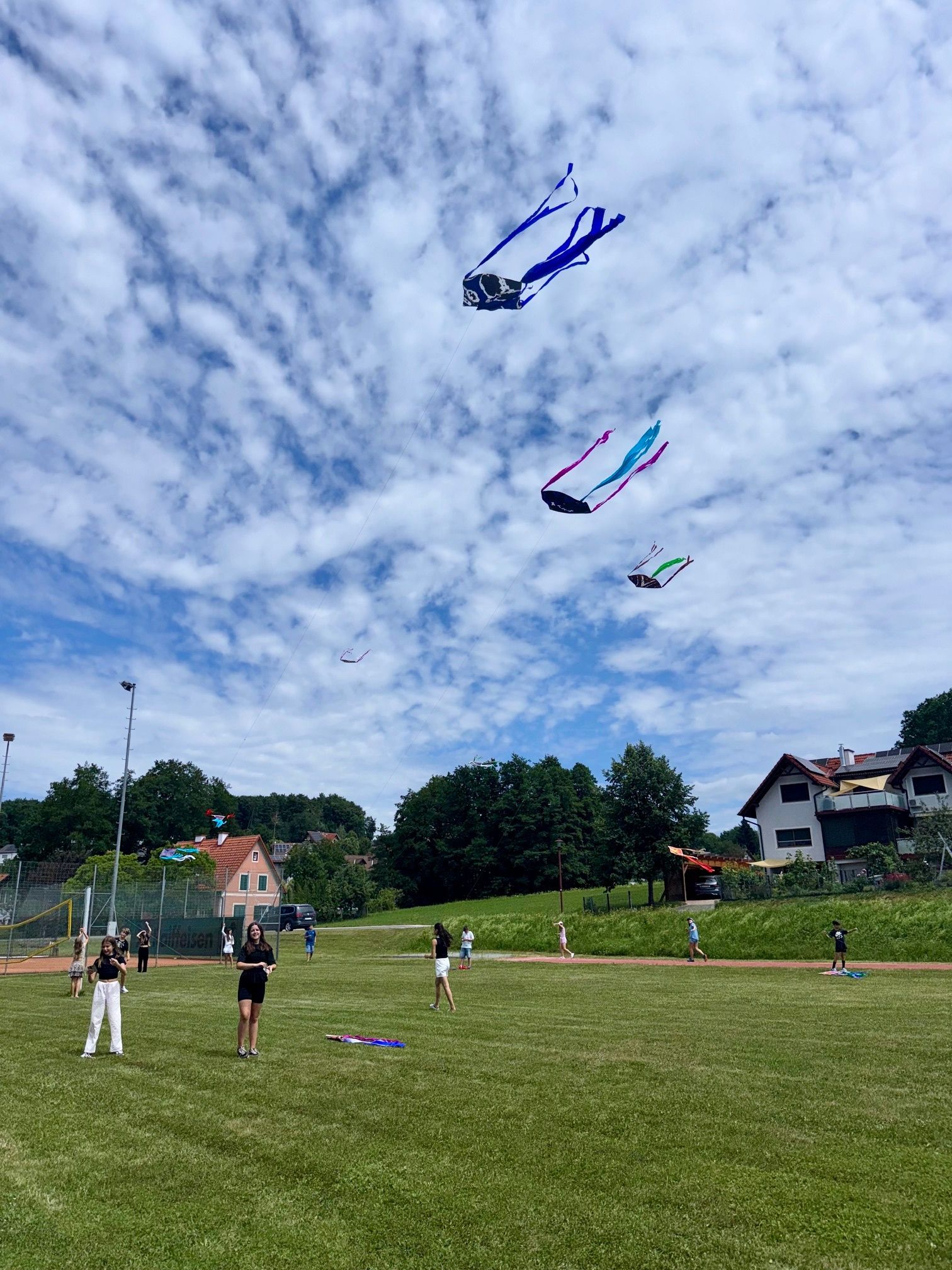 Menschen fliegen bunte Drachen auf einem Grasfeld. Ein Haus mit Solarpanelen befindet sich rechts. Bäume und Büsche umgeben das Gebiet. Der Himmel ist bewölkt.