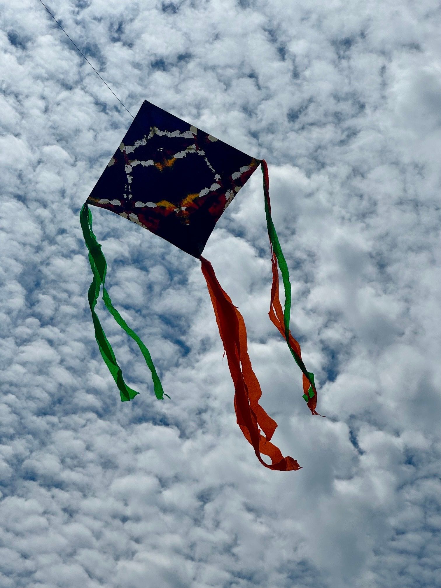 Ein diamantförmiges Drachen mit einem farbenfrohen Herzdesign fliegt in einem wolkenverhangenen blauen Himmel mit grünen und roten Schwänzen.
