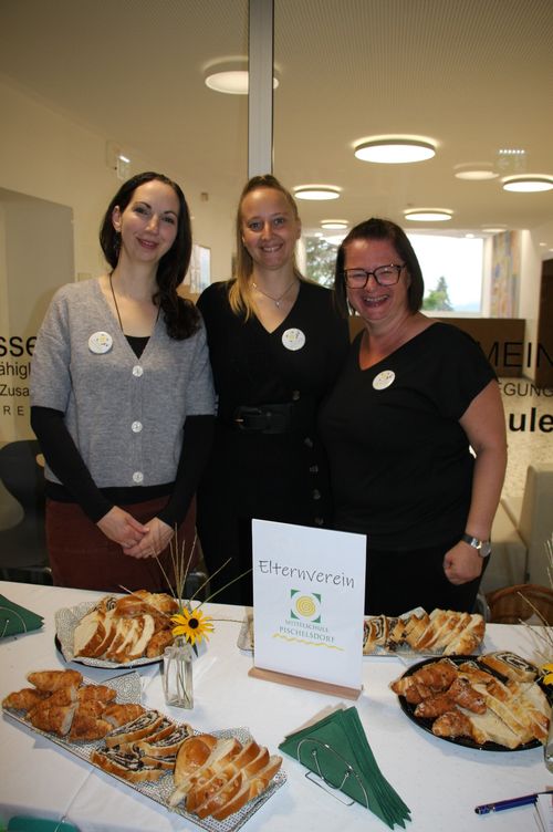 Drei Frauen stehen hinter einem Tisch mit einem Schild, das 'Elternverein' steht. Der Tisch ist mit verschiedenen Arten von Brot und Gebäck gefüllt.