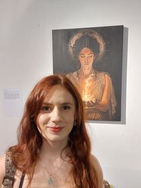 Eine Frau mit langen roten Haaren lächelt vor einem Gemälde einer Frau, die eine Flamme hält.