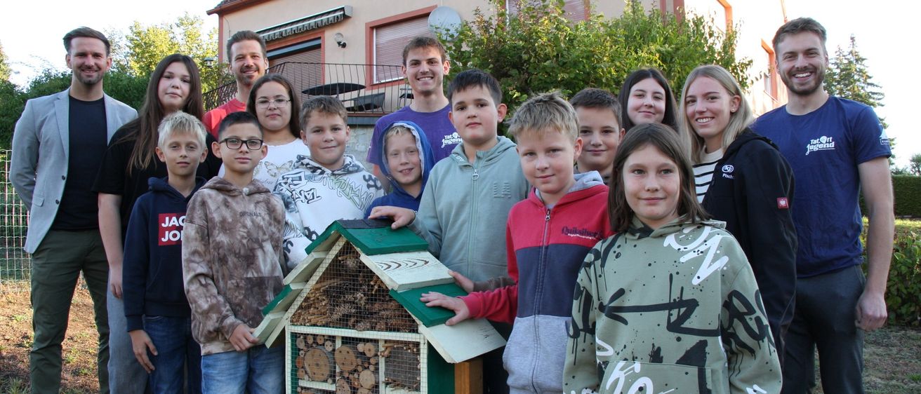 Eine Gruppe von Menschen, einschließlich Kindern, posiert für ein Foto vor einem Haus. Ein hölzerne Bienenhaus ist in der Mitte.