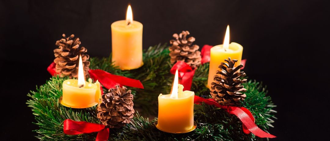 Ein Kranz aus Tannennadeln hält fünf brennende gelbe Kerzen, mit Tannenzapfen und roten Bändern. Die Kerzen und der Kranz stehen vor einem dunklen Hintergrund.