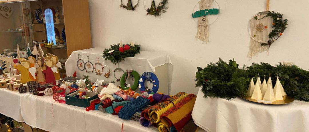 Ein Raum mit weißen Wänden und einem Bogengewölbe. Es gibt zwei Tische mit weißen Tischdecken. Ein Tisch hat verschiedene Gegenstände wie Schals, Mützen und Kränze. Ein anderer Tisch hat einen Kranz und einen Teller mit kleinen Zapfen. Die Wand hat drei hängende Dekorationen. Ein hölzerner Schrank mit Glastüren ist links.