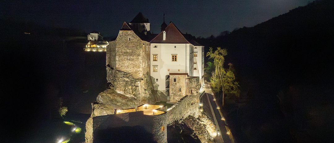 Eine mittelalterliche Burg auf einem felsigen Felsen ist bei Nacht beleuchtet, mit einem hellen Mond darüber. Die Burg hat Steinmauern und Türme und einen Weg, der zu ihr führt.