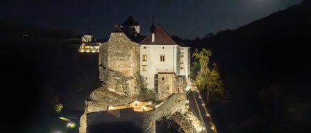 Eine Luftaufnahme einer Burg mit beleuchteten Wänden und Fenstern unter einem hellen Mond. Die Burg ist von Bäumen und Mauern umgeben, mit einem Pfad, der mit kleinen Lichtern beleuchtet ist.