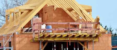 Ein Haus im Bau mit Holzrahmen und Ziegelwänden. Ein blauer Fass und verschiedene Baumaterialien befinden sich auf der Gerüstkonstruktion.