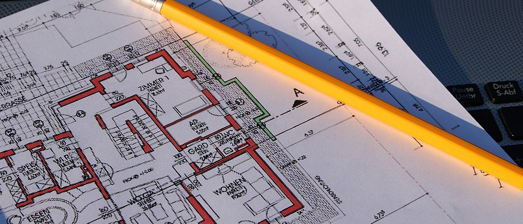 Ein gelber Bleistift liegt auf einem Bauplan mit architektonischen Details. Der Bauplan hat einen Grundriss mit Räumen, Fluren und Abmessungen. Der Radiergummi des Bleistifts ist oben links sichtbar.