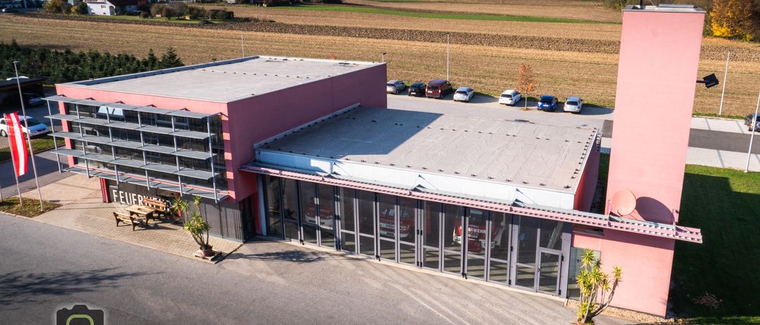 Luftaufnahme eines rosa Gebäudes mit Glaswänden, vor dem Autos geparkt sind, und einem großen Feld im Hintergrund.