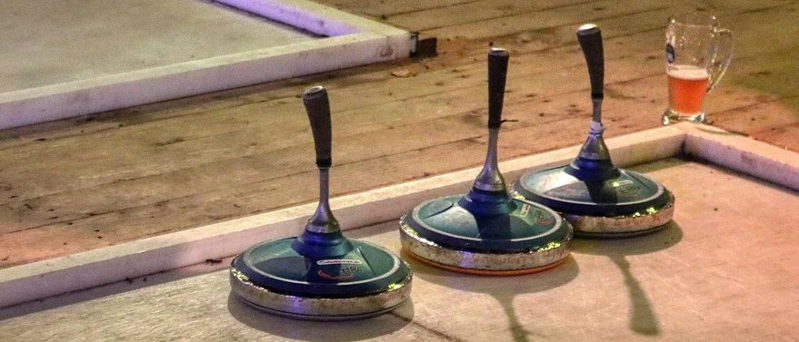 Drei blaue Curlingsteine stehen auf einem glatten Boden in der Nähe einer Holzkante. Schatten fallen auf den Boden. Ein Glas Bier steht an der Kante.