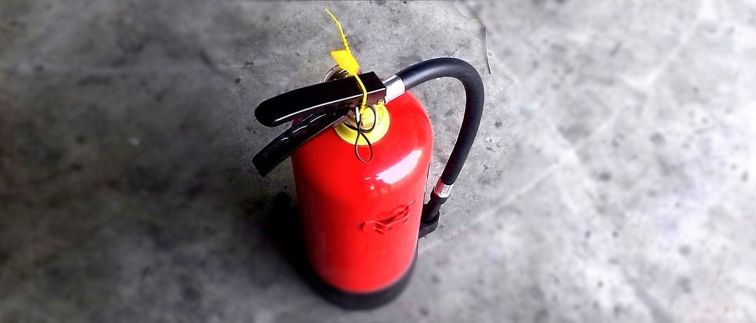 Ein roter Feuerlöscher mit einem gelben Etikett steht auf einem Betonboden. Er hat einen schwarzen Griff und eine schwarze Düse.