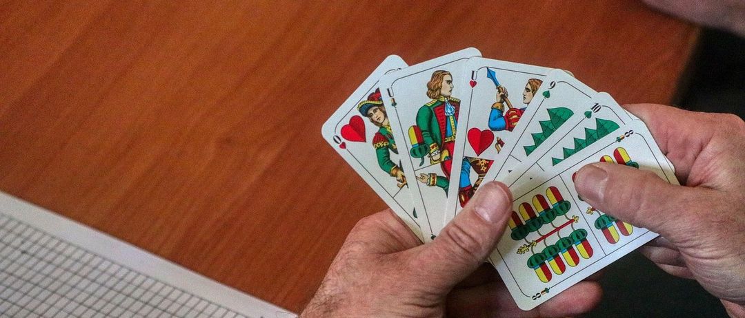 Eine Person hält eine Hand Karten auf einem Holztisch, ein Punktezettel liegt teilweise sichtbar in der Nähe.