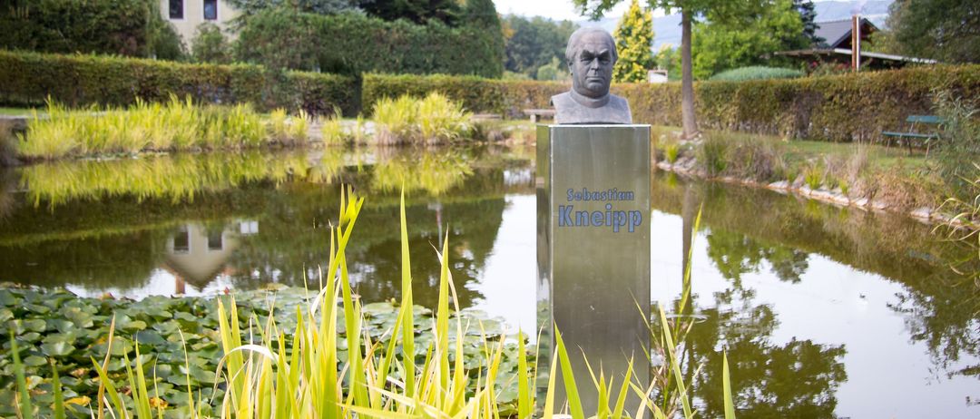 Eine Statue von Sebastian Kneipp befindet sich am Ufer eines Teiches. Die Statue ist aus Silber und hat den Namen 'Sebastian Kneipp' darauf. Die Umgebung ist üppig grün.