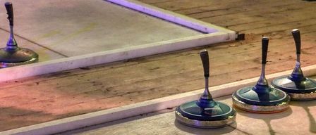 Three blue curling stones with handles are placed on a shiny floor. A wooden floor and a white edge are visible. A glass with a drink is placed on the edge.