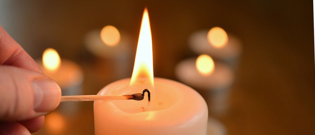 Eine brennende Kerze mit einem Streichholz in der Hand, umgeben von weiteren Kerzen im Hintergrund. Die Flamme ist hell und warm.
