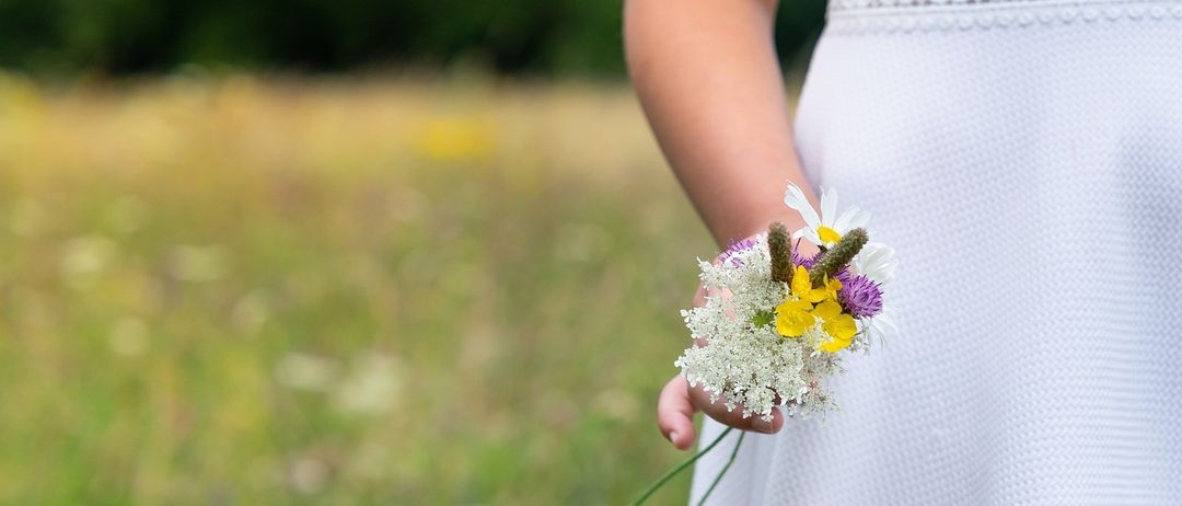 Eine Frau in einem weißen Kleid hält einen Blumenstrauß in einem Grasfeld. Der Strauß enthält verschiedene Blumen wie Gänseblümchen, lila und gelbe Blüten.