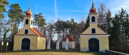 Zwei gelbe Kapellen mit Kupferkuppeln stehen vor einem Hintergrund von blauem Himmel und kahlen Bäumen. Ein großes Kreuz mit Statuen steht im Vordergrund, und eine kleinere Kapelle dahinter.