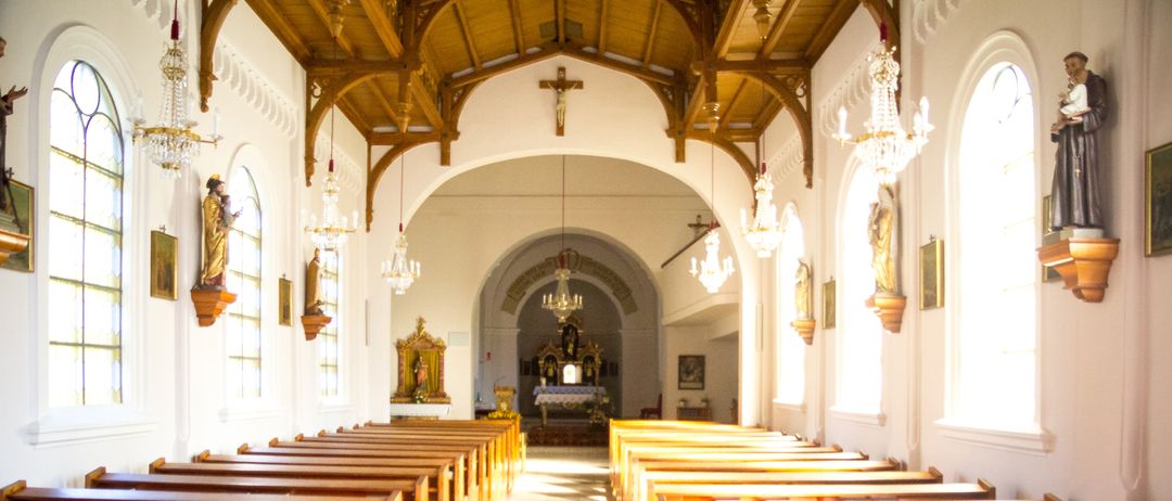 Ein leerer Kircheninnenraum mit hölzernen Kirchenbänken, einer hölzernen Decke und hängenden Kronleuchtern. Der Altar ist mit einem Kreuz geschmückt und von Statuen umgeben. Die Wände sind mit Bildern und Buntglasfenstern verziert.