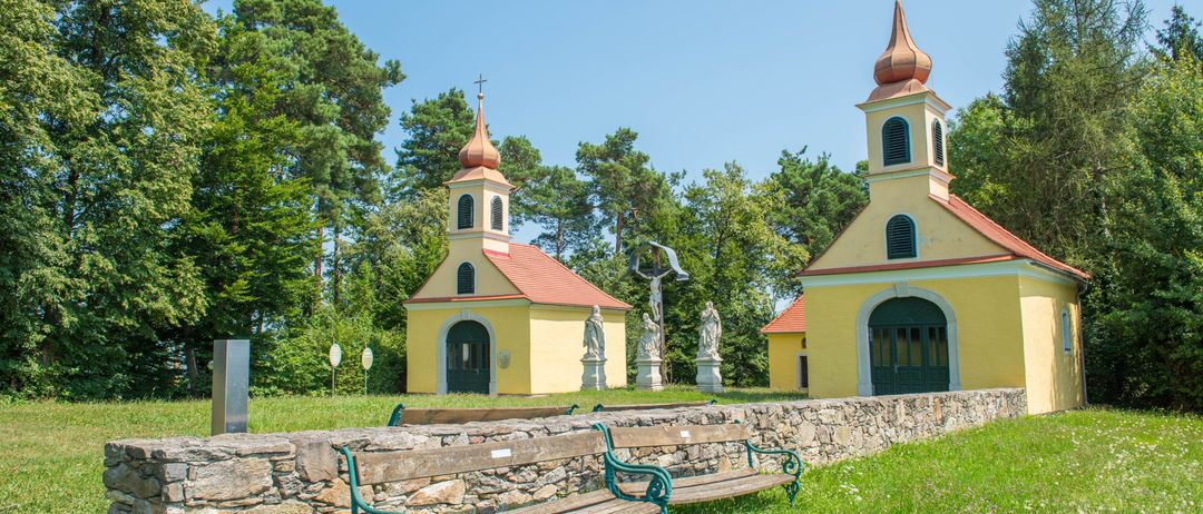 Eine gelbe Kapelle mit zwei Türmen, umgeben von Bäumen. Sie hat eine Steinmauer und eine Bank davor.