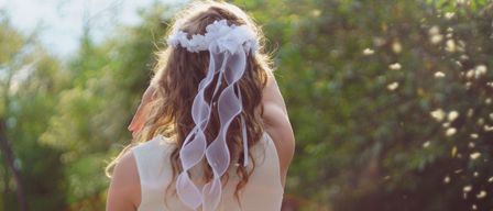 Ein Mädchen in einem weißen Kleid mit einer weißen Haarspange und Bändern schaut nach hinten, wobei Bäume im Hintergrund verschwommen sind.