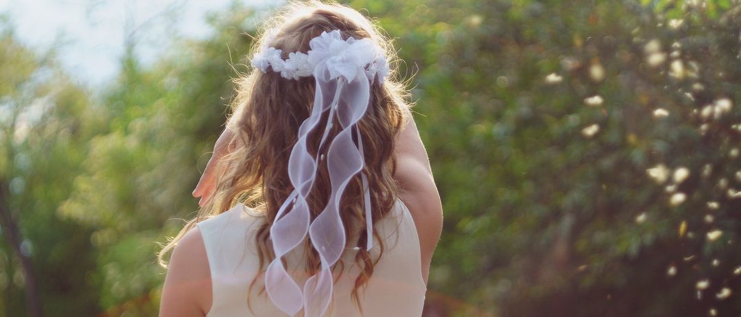 Ein Mädchen in einem weißen Kleid mit einer weißen Blumenkrone schaut nach links, mit Bäumen im Hintergrund.
