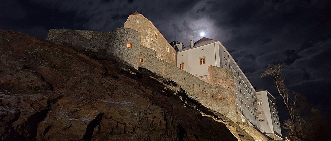 Eine Burg auf einem Felsen unter einem hellen Vollmond bei Nacht, beleuchtet vom Mondschein, mit sichtbaren Fenstern und einem Baum im Vordergrund.
