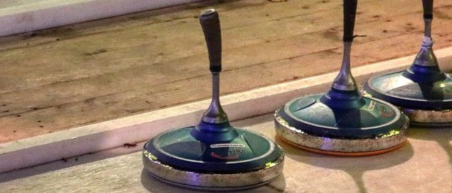 Drei Curlingbesen sind auf einem polierten Boden aufgereiht, wobei ein Schatten auf dem Boden geworfen wird. Im Hintergrund sind ein weißer Rand und eine hölzerne Oberfläche zu sehen.