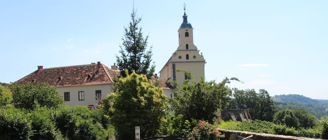Ein kleiner Garten mit gelben Blumen vor einer Kirche mit Turm. Ein Betonweg führt zu einer Bank.
