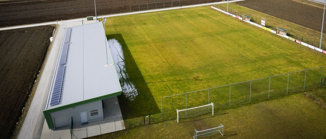 Eine Luftaufnahme eines Fußballfeldes mit einem Gebäude, das mit Solarmodulen ausgestattet ist und einem eingezäunten Bereich, umgeben von weitläufigen Feldern und einem Dorf in der Ferne.