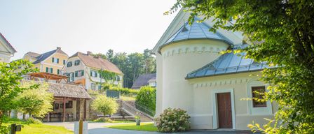 Ein Weg führt zu einem weißen Gebäude mit blauem Dach, umgeben von Grün. In der Nähe ist ein Haus mit braunem Dach und grünen Fensterläden zu sehen.
