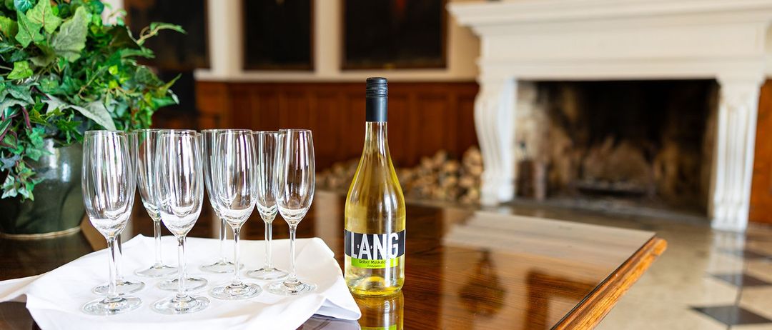 Eine Flasche Lang-Wein mit sechs leeren Weingläsern und einem weißen Tuch auf einem Holztisch in einem Raum mit Kamin und Bilderrahmen.