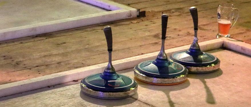 Drei Curlingbesen stehen auf einem polierten Holzboden. Schatten werden auf den Boden geworfen. Eine weiße Plattform und ein Holzboden sind im Hintergrund.