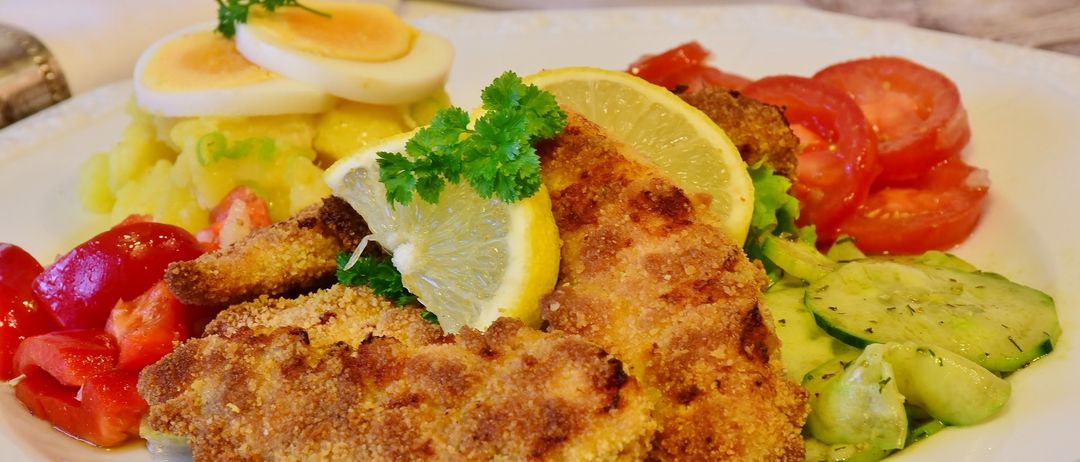 Ein weißer Teller mit gebratenem Fisch, garniert mit Zitrone und Petersilie. Dazu gelbe Kartoffelpüree, ein paar Tomatenscheiben und Gurken.