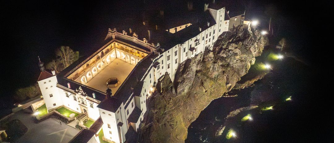 Ein Luftbild eines großen Schlosses auf einem Felsen, beleuchtet von Nachtlichtern. Das Schloss verfügt über einen zentralen Innenhof mit einem Brunnen, umgeben von Bogenfenstern und einem dunklen Himmelshintergrund.