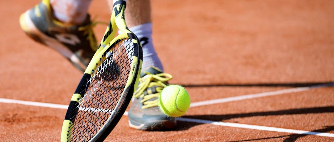 Ein Tennisspieler in Turnschuhen ist dabei, einen Tennisball auf einem Sandplatz zu schlagen. Der Ball ist gelb und der Schatten des Spielers wird auf den Boden geworfen.