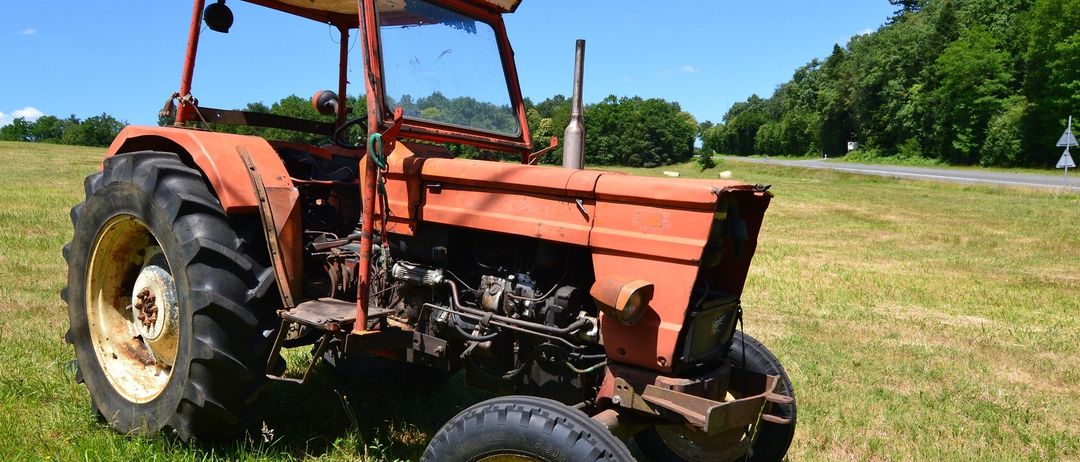 Ein alter roter Traktor mit dem Wort Sekura auf seiner Oberseite steht auf einem Grasfeld mit Blick auf eine Straße und Bäume in der Ferne.