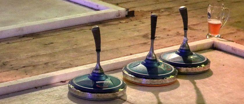 Drei Curlingbesen mit silbernen Griffen stehen auf einem polierten Holzboden. Sie sind eng beieinander positioniert, und ihre Schatten werden auf den Boden geworfen. Der Boden ist sauber und scheint gut gepflegt zu sein.