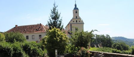 Ein friedlicher Garten mit gelben Blumen, einer Bank und einem Steinweg. Dahinter steht ein historisches Gebäude mit Turm, umgeben von üppiger Grünfläche.