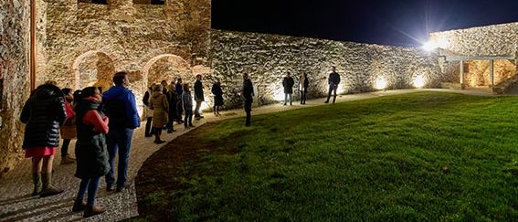 Touristen versammeln sich bei Nacht um eine mittelalterliche Burg, die von sanftem Licht erleuchtet wird. Die Burg hat Bogenfenster und eine Steinmauer.