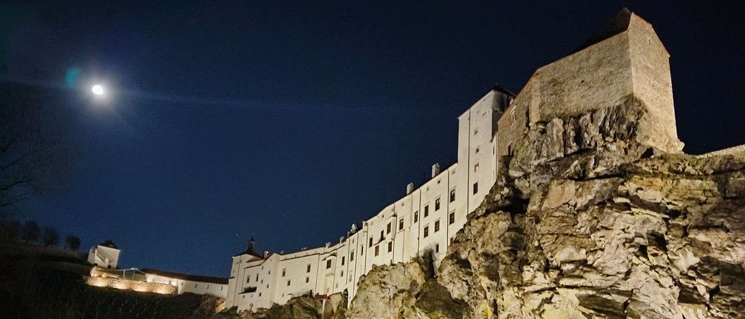 Eine Burg steht auf einem Felsvorsprung bei Nacht, beleuchtet von Lampen und dem hellen Mond.