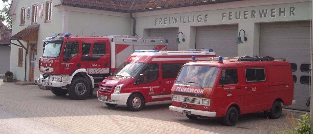 Three fire trucks are parked outside a fire station with the words Freiwillige Feuerwehr on the wall.