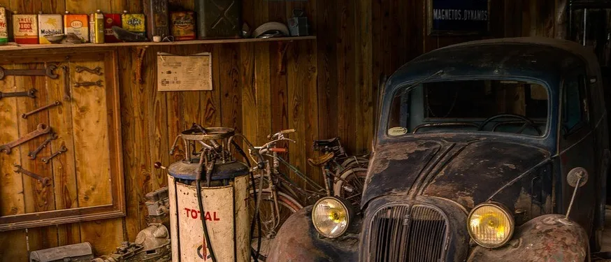Ein altes Auto mit Rost steht in einer Garage mit Holzwänden. Verschiedene Werkzeuge und Schilder sind an der Wand befestigt.