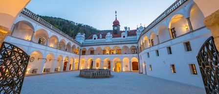 Ein großer Schlosshof, beleuchtet von warmen Lichtern, mit Bogenfenstern und einem Brunnen. Der Himmel ist blau mit einigen Wolken.