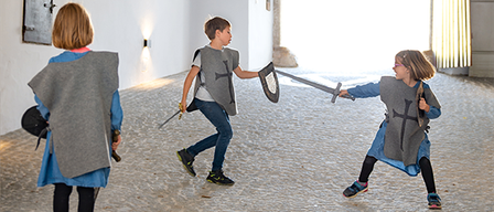 Drei Kinder, die als Ritter verkleidet sind, spielen mit Schwertern in einem Schlosskorridor. Sie schauen in Richtung Kamera.