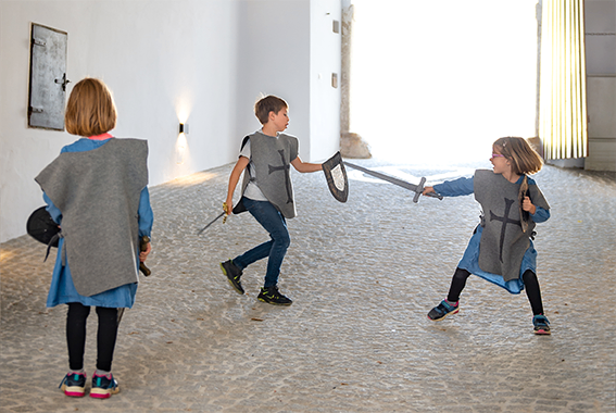 Drei Kinder, die als Ritter verkleidet sind, spielen mit Schwertern in einem Schlosskorridor. Sie schauen in Richtung Kamera.