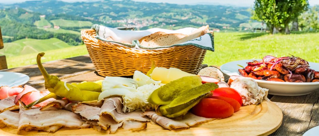 Ein Picknicktisch mit einer Auswahl an Fleisch, Käse, Gurken und Tomaten auf einem Holzbrett, mit einem Korb Brot im Hintergrund und einer malerischen Aussicht auf die Landschaft.