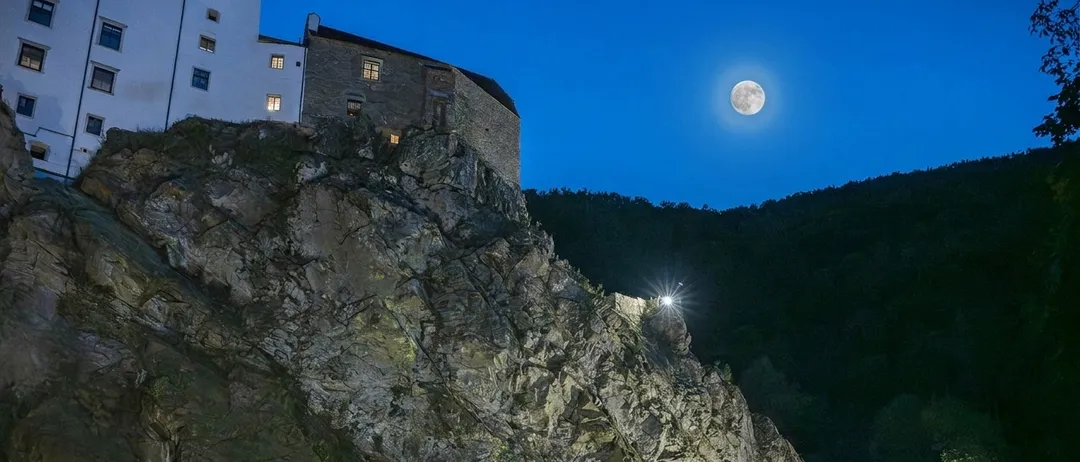 Eine Burg auf einem Felsvorsprung bei Vollmond in der Nacht, mit einem hellen Licht am Felsrand.
