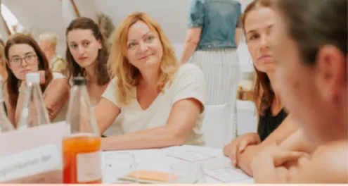 Eine Gruppe von Frauen sitzt an einem Tisch, möglicherweise in einer Besprechung. Eine Frau lächelt und schaut eine andere Frau an.
