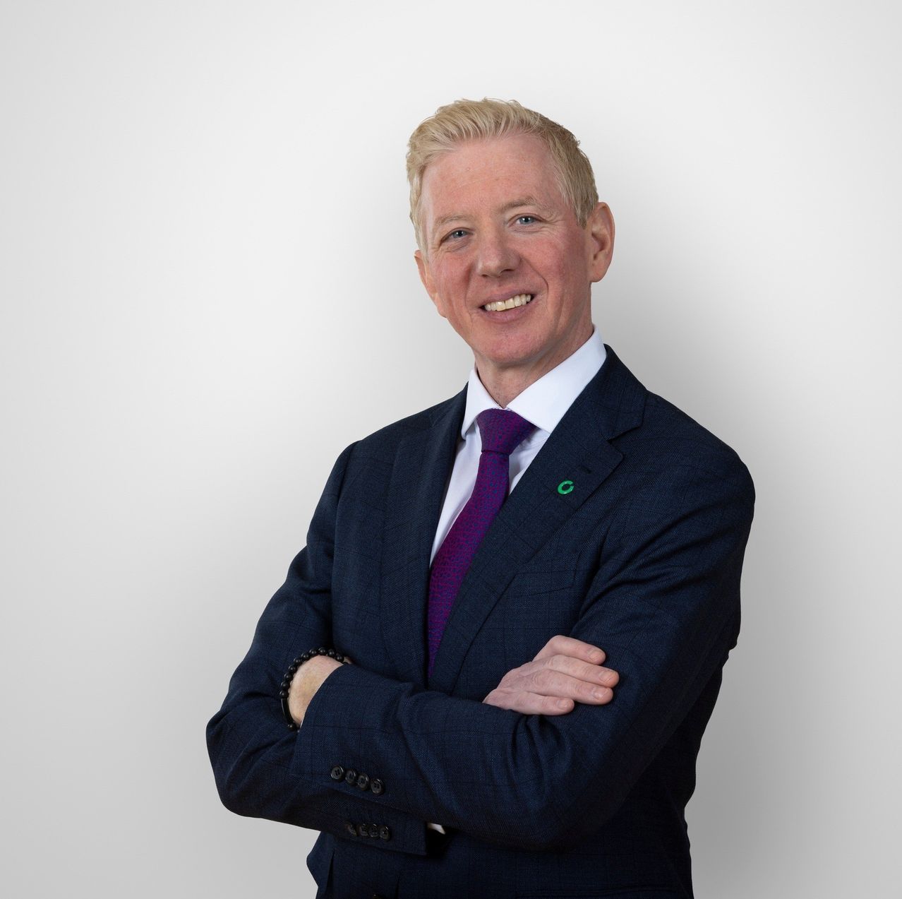 Ein Mann in einem dunklen Anzug und einer lila Krawatte lächelt und posiert für ein Foto mit verschränkten Armen.